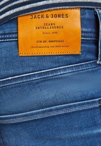 Blå denimjeans med en tan läderetikett med präglad text. Sticksömmen synlig längs fickorna och midjebandet.