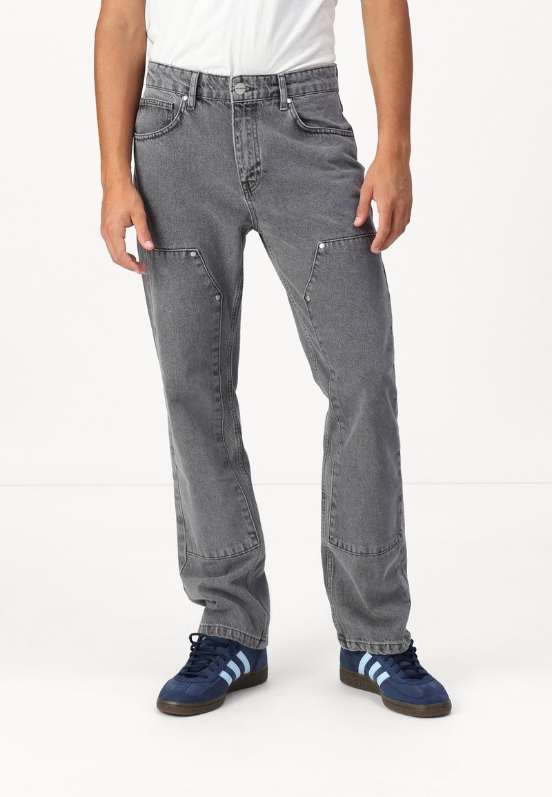 Jean en denim gris avec une coupe droite, présentant des coutures visibles, des poches latérales et des accents de quincaillerie argentée. Porté avec des chaussures de sport bleues.
