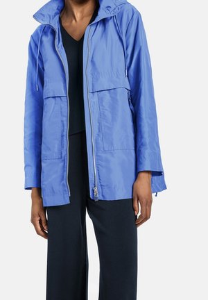 Personne portant une veste zippée bleu clair avec de grandes poches avant par-dessus un haut noir à col en V et un pantalon noir, debout devant un fond uni.