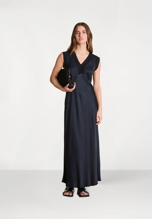 Femme debout en robe maxi noire sans manches, tenant une pochette noire, portant des sandales noires, sur un fond clair uni.
