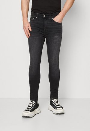 Homme portant un jean noir slim, des baskets montantes noires avec des semelles et des lacets blancs, debout devant un fond clair uni.