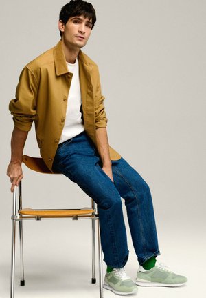 Een tan kleurige jack met knopen over een witte blouse, blauwe spijkerbroek en grijs sneakers met groene accenten. Gezeten op een houten stoel met een chroomframe.