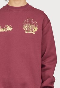 Sudadera burdeos con diseños gráficos dorados, que incluyen formas y figuras ornamentadas. Cuello redondo acanalado y corte relajado.