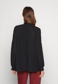 Blusa nera a maniche lunghe con dettagli arricciati su spalle e polsini, caratterizzata da un colletto alto e una silhouette fluida. Tessuto morbido e leggero.