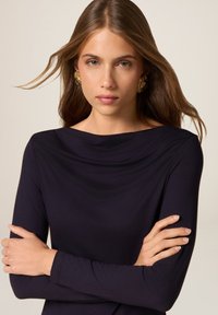 Donna con lunghi capelli castani, che indossa orecchini d'oro e un top nero a maniche lunghe, con le braccia incrociate, che guarda direttamente verso la fotocamera.