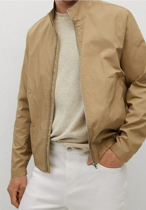 Homme portant une veste zippée marron clair sur un pull beige, avec un pantalon blanc, une main dans la poche, sur un fond uni.