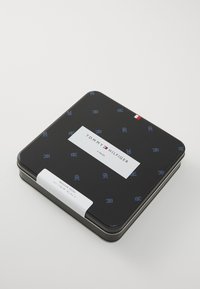 Schwarze Metalldose mit abgerundeten Ecken, geprägt mit Logo-Muster, weißes Etikett mit der Aufschrift "TOMMY HILFIGER", enthält vier Paare normale Socken.