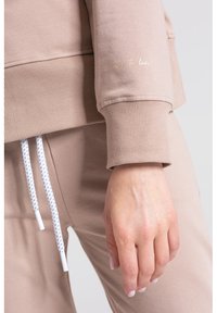 Beżowy materiał bluzy z ściągaczami, z białymi sznurkami. Ciekawy detal: "Łatwe do pokochania!" w złotym pisaniu na rękawie.