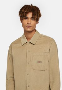 Beige ribfluwelen shirt met een klassieke kraag, knoopsluiting aan de voorkant en een patch pocket met logo-detail. Zachte textuur met verticale ribbels.