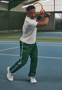 Pantaloni da track verdi con strisce bianche ai lati, T-shirt bianca e sneakers bianche, con una racchetta da tennis arancione in mano su un campo al coperto.
