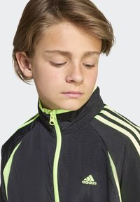 Crna sportska jakna s neonsko zelenim naglascima, visokim ovratnikom, patentnim zatvaračem i tri pruge na rukavima.