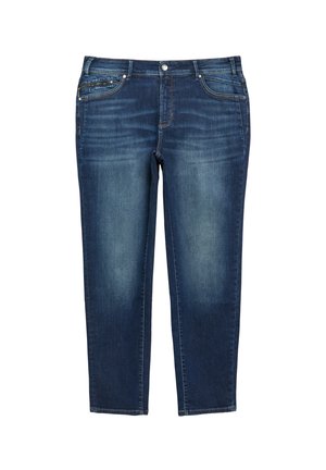 Frau steht mit einer Hand in der Tasche, trägt weiße Sneakers, dunkelblaue Jeans und ein weißes T-Shirt mit blauen horizontalen Streifen.