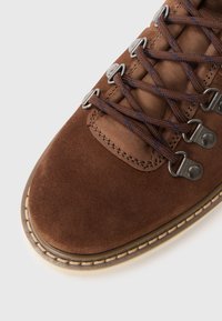 Chaussure de randonnée en daim marron avec œillets en métal, lacets marron, un col rembourré et une semelle en caoutchouc cousue. Vue rapprochée de l'orteil et du laçage.
