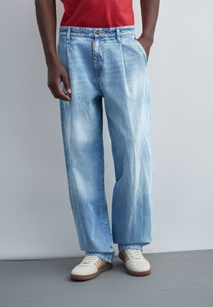 Muž nosi svetlomodré voľné rifle, biele tenisky s hnedými pruhmi a červenú košeľu, stojí s jednou rukou v vrecku.