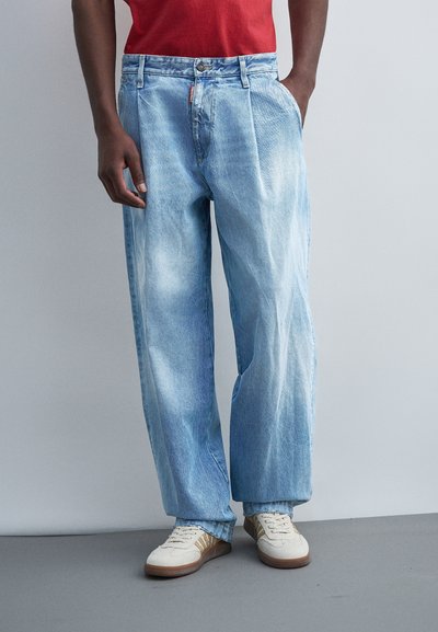 Homme portant un jean bleu clair ample, des baskets blanches avec des rayures marron et une chemise rouge, debout avec une main dans la poche.