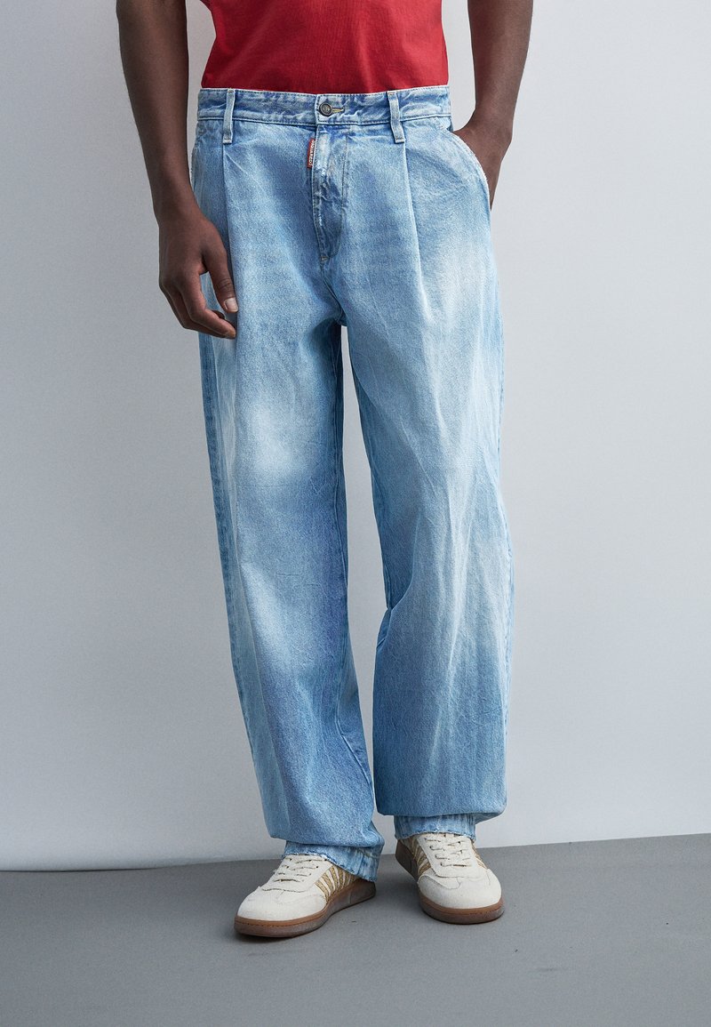 Άνδρας που φορά ανοιχτό μπλε άνετο τζιν, λευκά αθλητικά παπούτσια με καφέ ρίγες και κόκκινο πουκάμισο, στέκεται με το ένα χέρι στην τσέπη.