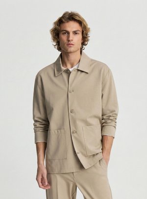 Giovane uomo con capelli biondi ricci che indossa una giacca beige con bottoni e grandi tasche e pantaloni abbinati, in piedi su uno sfondo bianco.