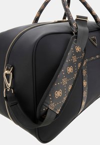Sac de sport noir avec une texture lisse, fermeture éclair et accessoires dorés, accentué par une sangle à motifs avec des motifs bruns sur fond noir.
