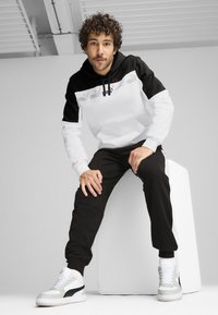 Crni i bijeli hoodie s grafičkim detaljima, uparen s crnim trenirkama i bijelim tenisicama. Kovrčava kosa, sjedeći položaj na svijetloj podlozi.