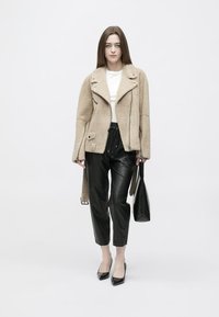 Vrouw draagt een beige shearling jas, witte top, zwarte leren broek, zwarte hakken, houdt een zwarte handtas vast, staand tegen een witte achtergrond.