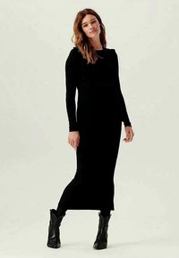 Vestido de jersey negro de manga larga con un cuerpo ajustado y corte recto. Llevado con botas negras hasta la rodilla. Textura suave, diseño minimalista.