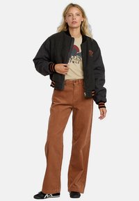 Zwart-grijze bomberjack met geribbelde manchetten, gecombineerd met brede bruine corduroy broek en een beige T-shirt met print.