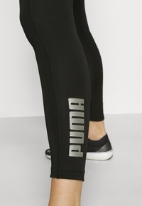 Czarne legginsy wykonane z gładkiego, elastycznego materiału. Posiadają wyraźne srebrne logo "PUMA" wzdłuż lewej strony oraz staranne detale szwów.