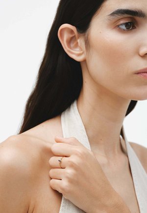 Anillo de oro con una pequeña piedra azul, llevado en una mano. Primer plano de la mano y el rostro, con piel suave y cabello oscuro. Diseño minimalista.