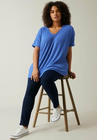 Blå, struktureret, kortærmet top med V-hals. Båret med mørke jeans og hvide sneakers, siddende på en træstol mod en neutral baggrund.