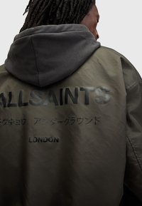 Khaki grøn bomberjakke med en grå hætte. Har fed sort tekst "ALLSAINTS" og "LONDON" samt glat, vandafvisende stof.