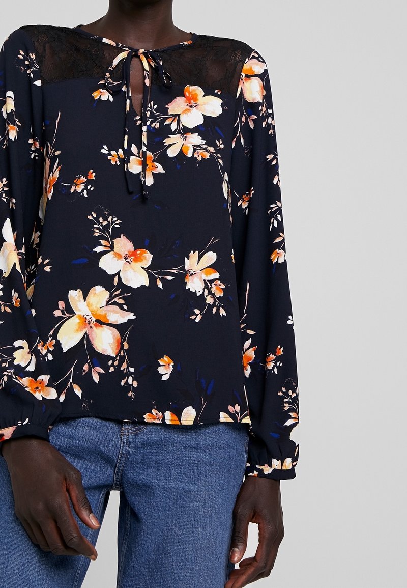 Blusa fluida blu navy con motivo floreale arancione e crema, yoke in pizzo e dettaglio con nodo al collo. Maniche lunghe con polsini elastici. Abbinata a jeans blu.