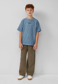 Blauwe t-shirt met korte mouwen en een ronde hals, voorzien van zwarte tekst, gecombineerd met losse olijfgroene broeken en witte sneakers met lichtbruine zolen.