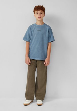 Blauwe t-shirt met korte mouwen en een ronde hals, voorzien van zwarte tekst, gecombineerd met losse olijfgroene broeken en witte sneakers met lichtbruine zolen.