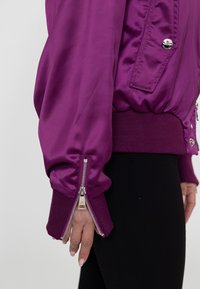 Paarse satijnen bomberjack met geribde manchetten en ritssluitingen op de mouwen, voorzien van een ronde hals en metalen hardwaredetails.