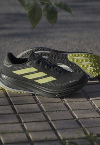 Czarne buty biegowe z materiałem GORE-TEX, żółtymi akcentami, teksturowaną powierzchnią i solidnym bieżnikiem, wystawione na nawierzchni asfaltowej.