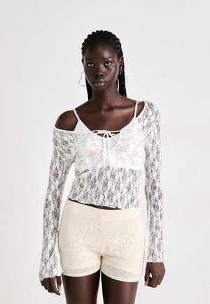White lace blouse with floral patterns, off-shoulder design, long bell sleeves, paired with cream crochet shorts featuring textured accents.