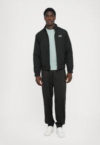 Black zip-up jacket with a high collar, featuring a logo on the chest, paired with a light blue shirt and black trousers. White shoes.