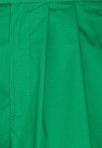 Zelená látka s sklady, která předvádí hladkou texturu a rovnoměrné švy. Viditelná boční kapsa zvýrazňuje funkčnost.