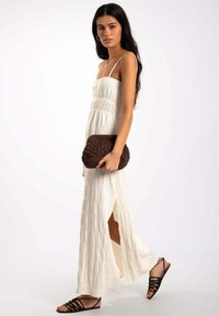 Robe maxi blanche texturée avec des fines bretelles, des fentes latérales et une taille froncée. Associée à une pochette en tricot marron et des sandales noires à lanières.