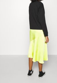 Sort langærmet top, parret med en lys gul asymmetrisk nederdel. Nederdelen har en silkeagtig tekstur, og outfittet fuldendes med sorte sneakers.