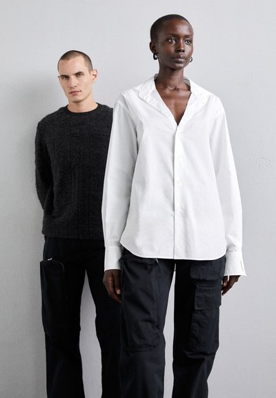 A man in a dark textured sweater stands behind a woman in a white button-up shirt and black cargo pants with visible pockets.