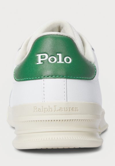 Vit sneaker med en grön läderhälla. Har vita snören, präglad branding och en texturerad gummisula. Ren design, minimala accentfärger.