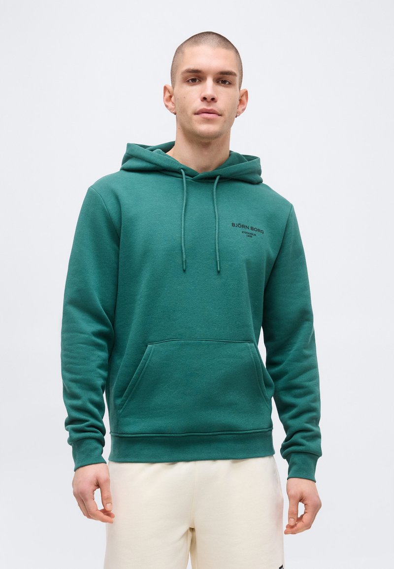 Ung mann med kort hår iført en grønn Björn Borg-hettegenser og lyse bukser, stående foran en ensfarget hvit bakgrunn.