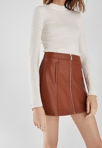 Hnědá kožená mini sukně s předním zipem, se strukturovanými sklady a bočními kapsami, spárovaná s přiléhavým bílým žebrovaným topem s dlouhým rukávem.