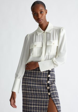 Blusa bianca con due tasche sul petto e chiusura a bottoni, abbinata a una gonna a quadratini blu navy e crema con bottoni laterali e uno spacco.