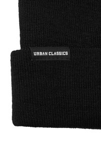 Bonnet en tricot noir avec un revers, arborant une étiquette rectangulaire portant le texte "URBAN CLASSICS" en lettres blanches. Texture douce et motif côtelé.