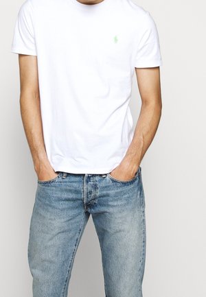 Biała bawełniana koszulka z okrągłym dekoltem i małym zielonym logo haftowanym na piersi, w połączeniu z jasnoniebieskimi dżinsami.