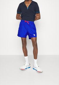 Polo a maniche corte blu navy con colletto a zip; pantaloni corti blu brillante con cordoncino rosso; scarpe da ginnastica bianche con accenti rossi e blu; vestibilità casual.