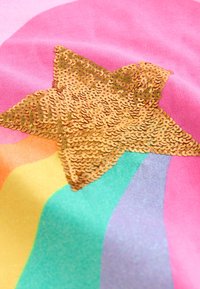 Rožnata tkanina ima veliko zlato bleščečo zvezdo na mavričnem vzorcu z zelenimi, modrimi, rumenimi in vijoličnimi črtami.