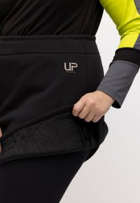 Zwarte sportrok met een zachte, geweven binnenvoering. Bevat een logo "UP SPORTS" en een rekbare tailleband. Het model houdt de zoom vast.